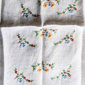 May include: White linen fabric with embroidered floral patterns. The embroidery features blue, yellow, and red flowers with green leaves. The fabric has a textured appearance and a green border.