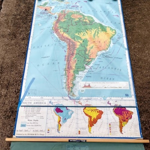 May include: A vintage, rolled-up map of South America with a blue background and green, yellow, and brown landmasses. The map includes a key with information about water depths, cities, and land elevation. The map is titled "South America" and is published by A.J. Nystrom & Co., Chicago.