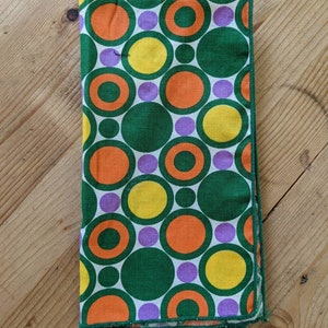May include: Folded green, orange, yellow, and purple patterned fabric. The fabric features a repeating pattern of concentric circles in various sizes and colors. The fabric is folded neatly and appears to be a napkin or a similar textile item.