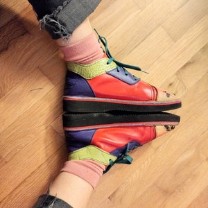 Puede incluir: Un par de zapatillas coloridas con un dise&ntilde;o multicolor, que incluye rojo, verde, amarillo y morado. Las zapatillas se llevan con calcetines y pantalones vaqueros.
