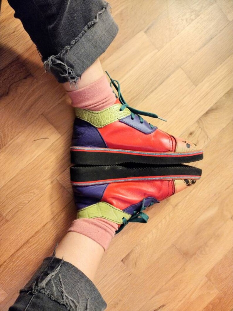 Puede incluir: Un par de zapatillas coloridas con un dise&ntilde;o multicolor, que incluye rojo, verde, amarillo y morado. Las zapatillas se llevan con calcetines y pantalones vaqueros.