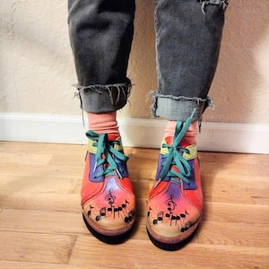 Puede incluir: Un par de zapatos rojos, naranja, amarillos y morados con notas musicales negras pintadas en ellos. Los zapatos tienen cordones verdes y los lleva una persona que lleva pantalones vaqueros azules.