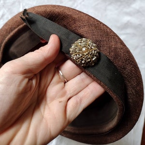 Könnte beinhalten: Ein brauner Hut mit einem dunkelgrünen Band und einem dekorativen, perlenbesetzten Schmuck. Der Hut wird von einer Hand gehalten, die die Textur des Stoffes und die komplizierten Details des Schmucks zeigt. Das Design des Hutes deutet auf einen Vintage- oder Retro-Stil hin.