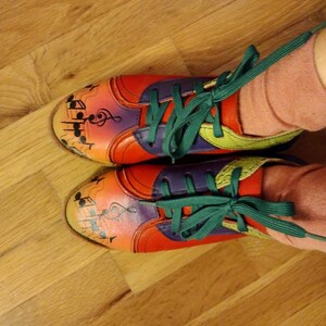 Puede incluir: Un par de zapatillas coloridas con notas musicales y una clave de sol pintadas en ellas. Las zapatillas tienen cordones verdes y est&aacute;n atadas con un lazo. Las zapatillas est&aacute;n sobre un suelo de madera.