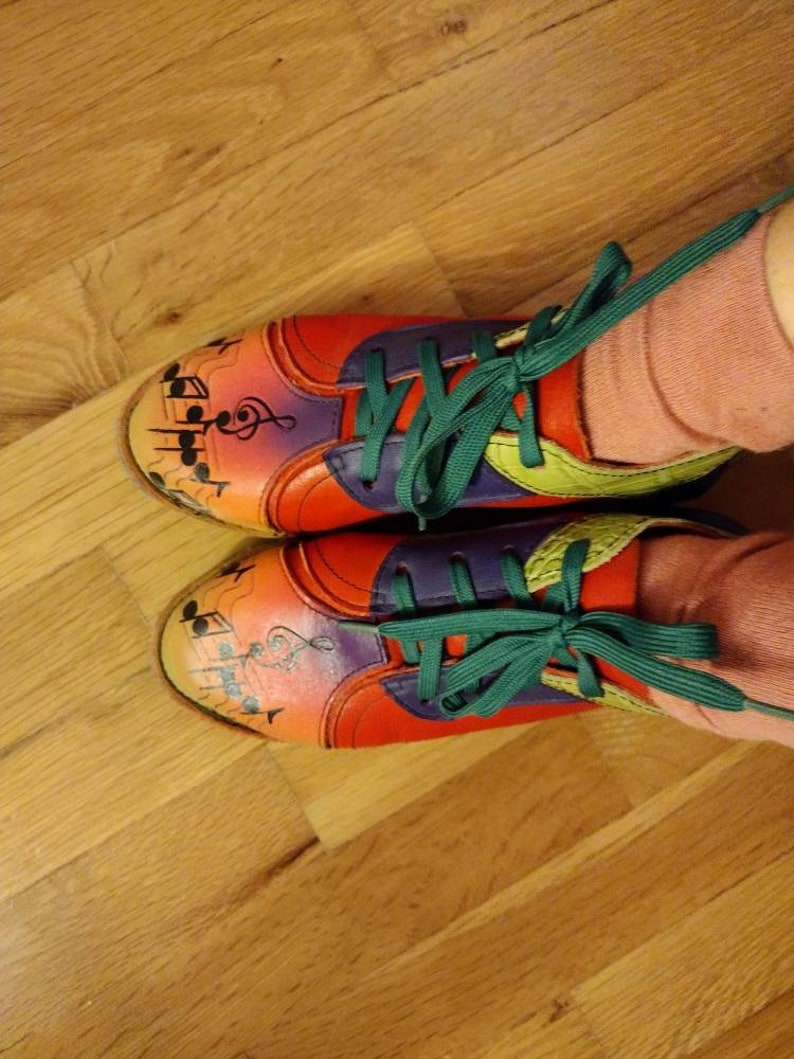 Puede incluir: Un par de zapatillas coloridas con notas musicales y una clave de sol pintadas en ellas. Las zapatillas tienen cordones verdes y est&aacute;n atadas con un lazo. Las zapatillas est&aacute;n sobre un suelo de madera.