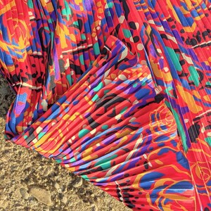May include: Red pleated fabric with a colourful abstract floral pattern. The fabric has a silky sheen and is folded in a way that shows the pleats.