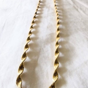Può includere: Una collana color oro con un design a corda intrecciata. La collana è realizzata in un materiale metallico lucido ed è esposta su una superficie bianca. La catena è lunga circa 45 cm. Questo gioiello è adatto a varie occasioni.