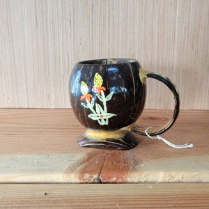 May include: A small, hand-painted coconut shell mug with a brown exterior and a yellow base. The mug features a floral design with orange and yellow flowers and green leaves.