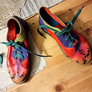Puede incluir: Un par de zapatos de cuero coloridos con cordones verdes. Los zapatos tienen un dise&ntilde;o rojo, morado y verde con notas musicales pintadas en los dedos.