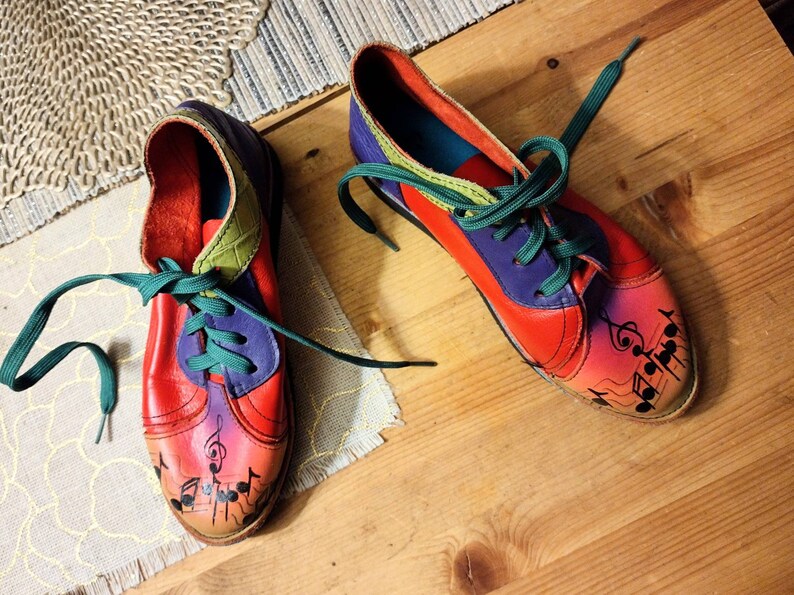 Puede incluir: Un par de zapatos de cuero coloridos con cordones verdes. Los zapatos tienen un dise&ntilde;o rojo, morado y verde con notas musicales pintadas en los dedos.