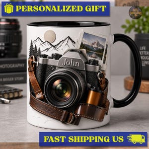 Peut inclure: Un mug en céramique blanc avec une anse et un bord noirs, orné d'un appareil photo vintage avec le prénom John imprimé sur le devant. Le motif comprend une sangle en cuir marron, une pellicule photo et un paysage de montagne avec un soleil.