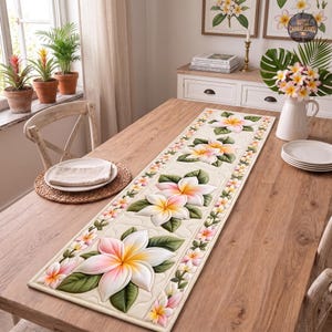 Puede incluir: Un camino de mesa rectangular de tela acolchada color crema, decorado con grandes flores de plumeria impresas con aspecto tridimensional en tonos blanco, rosa y amarillo, rodeadas de hojas verdes y un borde de flores más pequeñas.