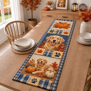 May include: A rectangular quilted table runner featuring a blue and white plaid border. The design includes panels with golden retriever dogs, orange pumpkins, autumn maple leaves, and dark blue paw prints on a cream-coloured background.