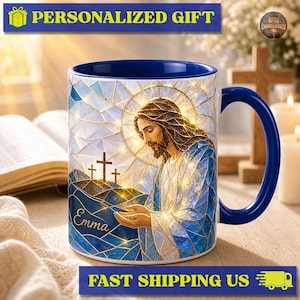 May include: A ceramic mug with a dark blue interior and handle, featuring a stained-glass style illustration of a bearded figure with long hair, hands cupped, and three crosses on a hill. The name Emma is printed in gold script on the side.
