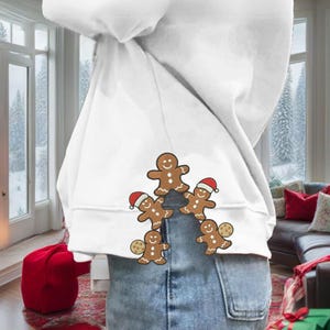 Może przedstawiać: Biała bluza z kapturem z motywem pięciu piernikowych ludzików. Czterech piernikowych ludzików ma czerwone czapki Świętego Mikołaja, a jeden trzyma ciasteczko. Wzór znajduje się na dolnej części pleców. Osoba ma na sobie dżinsy.