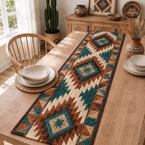 May include: A rectangular quilted table runner featuring a geometric pattern in shades of teal, brown, cream, and dark orange. The fabric displays repeating diamond and zigzag motifs with visible stitching throughout the length of the runner.