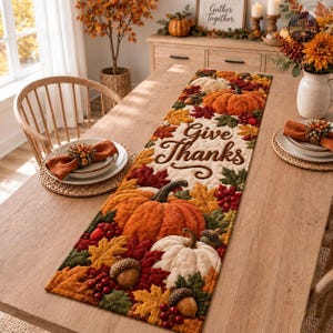 Op de afbeelding: Een rechthoekige, doorgestikte tafelloper met een herfstpatroon van oranje en witte pompoenen, rode bessen, eikels en kleurrijke herfstbladeren. De tekst "Give Thanks" is in bruin cursief schrift in het midden van de loper geborduurd.