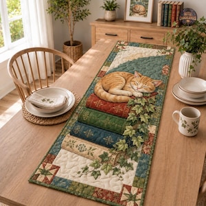 Può includere: Un runner da tavola trapuntato rettangolare con un disegno stampato di un gatto soriano arancione che dorme su una pila di quattro libri antichi. Il runner è decorato con viticci di edera verde e un bordo patchwork nei toni crema, verde e rosso.