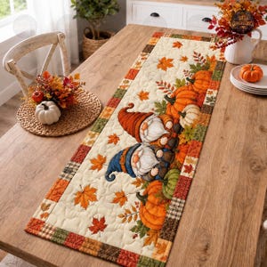 Può includere: Un runner da tavola trapuntato rettangolare con sfondo color crema, decorato con due gnomi, zucche arancioni e foglie d'acero autunnali. Il bordo presenta un motivo patchwork nei toni del marrone, dell'arancione e del verde.