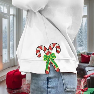 May include: White sweatshirt featuring a Christmas candy cane design. The candy canes are red and white with a green bow. The design is embroidered on the back of the sweatshirt.