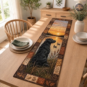May include: A rectangular table runner featuring a central scene of a black Labrador retriever sitting in tall grass by a lake at sunset, with two ducks swimming nearby. The runner is bordered by a patchwork pattern of brown and orange autumn leaves.