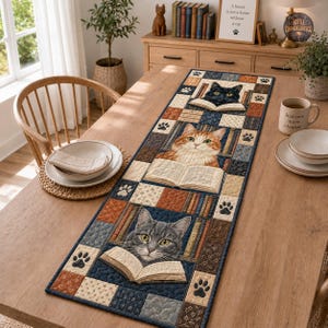 May include: A rectangular quilted table runner featuring a patchwork design with three cats reading books, surrounded by bookshelves and paw print motifs. The color palette includes shades of blue, brown, cream, and orange, with detailed cat illustrations.