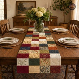 May include: A rectangular quilted table runner featuring a patchwork design of various fabric squares in shades of burgundy, navy blue, olive green, cream, and floral patterns, centered on a wooden dining table with place settings and flowers.