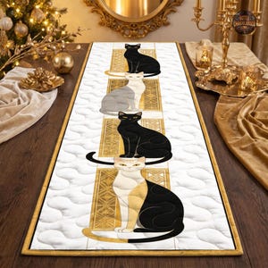 Peut inclure: Un chemin de table rectangulaire doté d'une surface matelassée blanche avec un motif répétitif de quatre chats dans des tons noir, blanc et or. Chaque chat est placé dans un cadre rectangulaire doré vertical avec des détails géométriques.