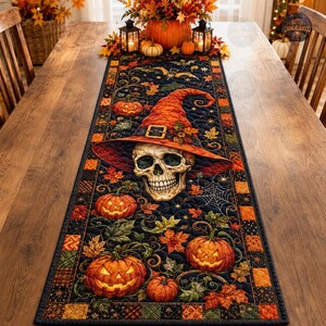 May include: A rectangular quilted table runner featuring a dark blue background with a central human skull wearing a large orange witch hat. The design includes multiple jack-o'-lanterns, autumn leaves, spider webs, and a chequered border.