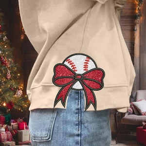 Op de afbeelding: Een beige hoodie met een rode en witte baseball met een rode strik erop. De hoodie wordt door iemand gedragen en de strik zit op de achterkant van de hoodie.