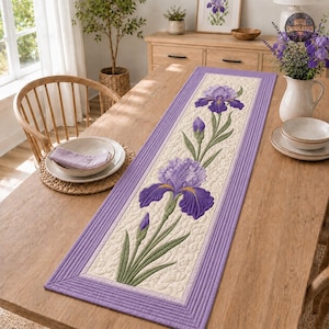 May include: A rectangular quilted table runner featuring a cream-colored center panel with embroidered purple iris flowers and green stems. The runner has a wide lavender border with parallel stitched lines and a textured, wavy quilted pattern.