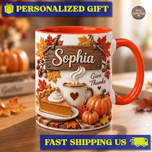 May include: A white ceramic mug with an orange interior and handle, featuring a 3D-style autumn design with a wooden sign reading Sophia, a slice of pumpkin pie, a small mug of coffee, pumpkins, and fall leaves with the text Give Thanks.