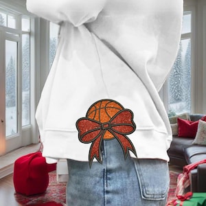 May include: A white sweatshirt with a red and orange basketball with a red bow on the back. The sweatshirt is being worn by a person with blue jeans.