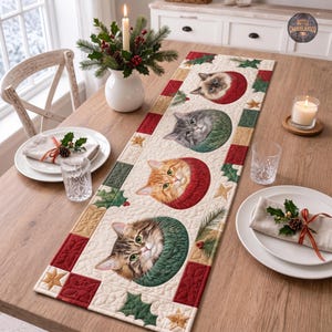 May include: A rectangular quilted table runner featuring four cat faces peeking out from colourful knitted sweaters. The runner has a cream background with red and green borders, decorated with holly leaves, berries, and gold star patterns.