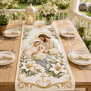 Pode incluir: Um caminho de mesa cor de creme com um design em relevo da Sagrada Família com o menino Jesus, rodeado por lírios brancos e volutas douradas. O caminho está centrado numa mesa de madeira com talheres e uma lanterna.
