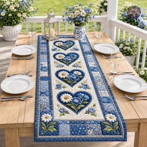 May include: A rectangular quilted table runner featuring a repeating pattern of blue and white heart-shaped floral motifs with daisies and blue blossoms. The runner has a blue and white patchwork border and is set on a wooden table with plates.