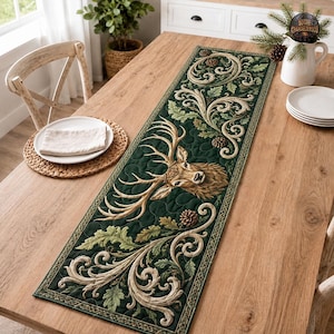 May include: A rectangular table runner featuring a dark green quilted fabric with a central stag head illustration, surrounded by intricate beige scrollwork, oak leaves, and pinecones. The runner has a decorative border and a stitched texture.