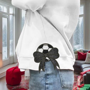 Könnte beinhalten: Weißer Kapuzenpullover mit einem schwarz-weißen Fußball-Design und einer schwarzen Schleife. Der Kapuzenpullover wird mit einer blauen Jeans getragen. Das Design ist auf der Rückseite aufgestickt. Der Hintergrund umfasst ein Fenster und einen roten Hocker.