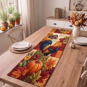 May include: A rectangular fabric table runner featuring a patchwork design with a central rooster, orange pumpkins, and autumn leaves in shades of red, yellow, and green. The runner is laid flat on a wooden table, showcasing a detailed seasonal print.