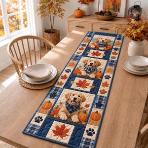 May include: A rectangular fabric table runner featuring a repeating pattern of golden retriever puppies wearing blue plaid bandanas, orange pumpkins, dark blue paw prints, and autumn maple leaves in shades of red, orange, and yellow on a blue background.