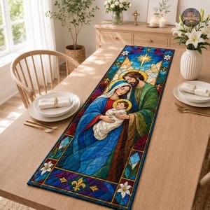 Pode incluir: Um caminho de mesa retangular com uma estampa estilo vitral da Sagrada Família. O design mostra uma figura central segurando um bebê, cercada por padrões geométricos azuis, verdes e roxos com detalhes de lírios brancos e uma estrela.