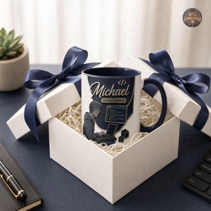 Peut inclure: Un mug en c&eacute;ramique blanche avec un int&eacute;rieur et une anse bleu fonc&eacute;, orn&eacute; d'un graphique repr&eacute;sentant une personne travaillant sur un ordinateur, le nom Michael, le texte PROGRAMMER et des symboles de code, pr&eacute;sent&eacute; dans une bo&icirc;te cadeau.