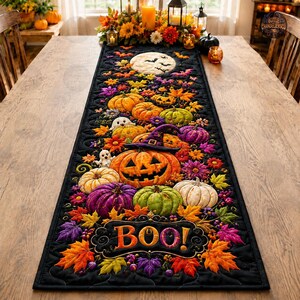 Puede incluir: Un camino de mesa acolchado negro con temática de Halloween que presenta una calabaza central grande con un sombrero de bruja morado, rodeada de hojas de otoño coloridas, fantasmas pequeños, calabazas de varios colores y una luna llena con el texto BOO.
