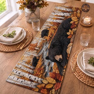 Peut inclure: Un chemin de table matelassé rectangulaire présentant une scène imprimée d'un grand ours noir et de deux oursons dans une forêt automnale avec des bouleaux et des feuilles orange, jaunes et brunes. Le chemin a une texture piquée.
