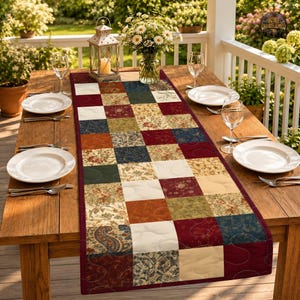 May include: A rectangular quilted table runner featuring a patchwork design of squares in shades of burgundy, navy blue, olive green, cream, and tan. The fabric squares display various floral, paisley, and solid patterns with visible stitching.