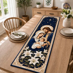 Pode incluir: Um caminho de mesa retangular com fundo azul escuro e textura acolchoada. O design apresenta uma cena religiosa de uma família com um bebé, rodeados por lírios brancos e detalhes dourados, emoldurados por uma borda decorativa cor de creme.