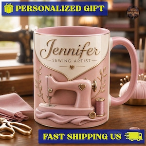 Peut inclure: Un mug en céramique aux tons rose et crème avec un relief représentant une machine à coudre, une bobine de fil et un bouton. Le texte Jennifer et Sewing Artist est imprimé sur le devant dans une police dorée de style script et serif.