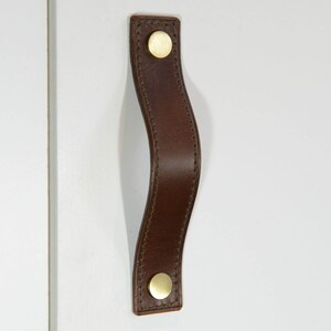 May include: A brown leather cabinet pull with a curved shape and two gold metal knobs.