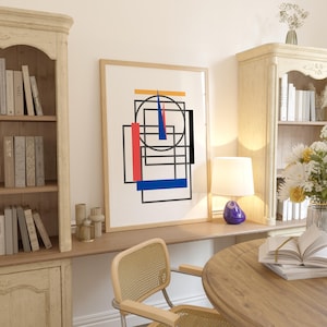 May include: A framed abstract art print featuring geometric shapes in red, blue, black, and yellow. The artwork is displayed in a light-colored wooden frame. The print is set in a room with a desk, chair, and bookshelves.