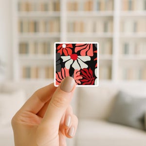 May include: A square sticker featuring a floral pattern in red, white, and black. The design includes stylized flowers and leaves against a black background. The sticker is held by a hand.